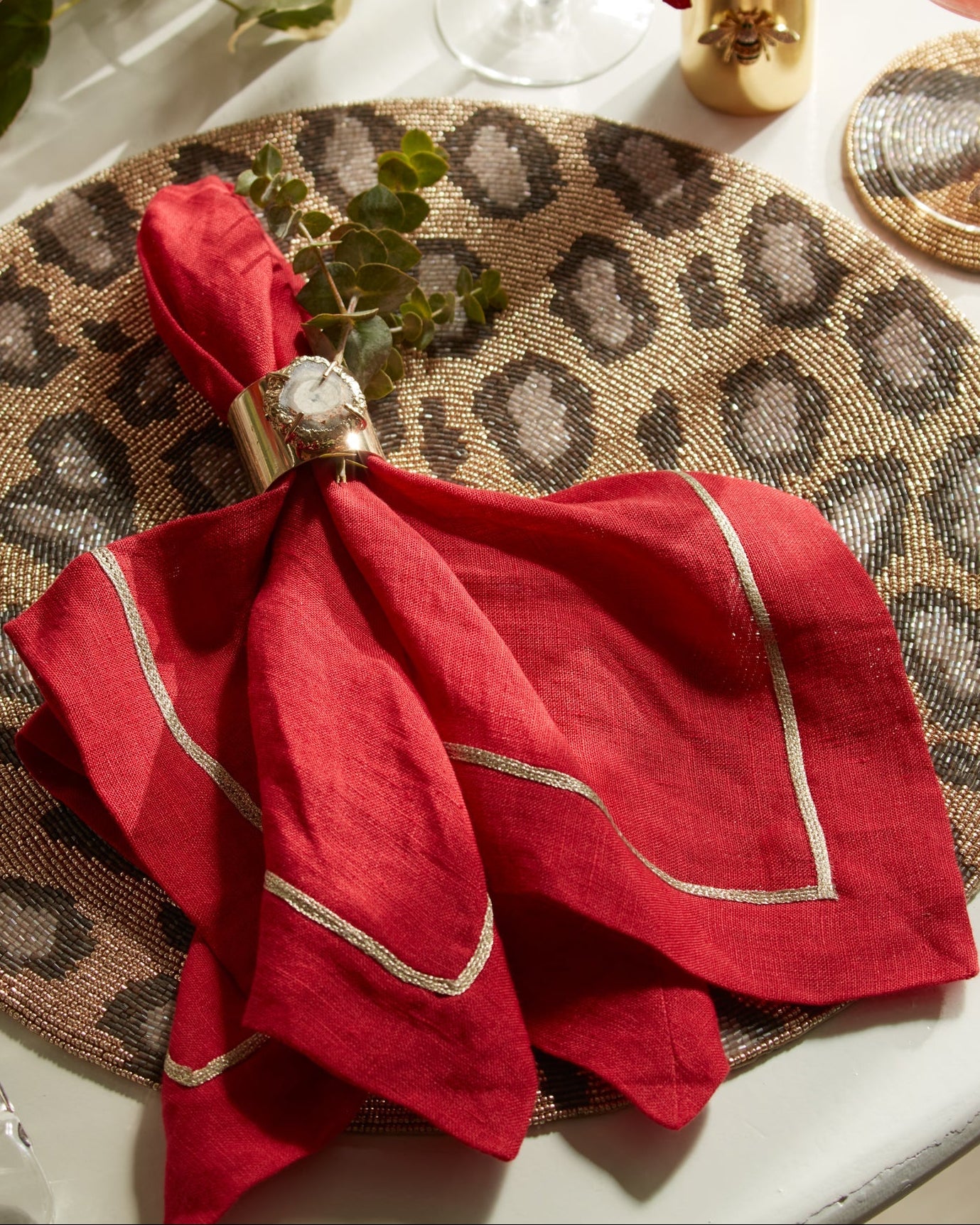 Red napkin with gold trim on a leopard print placemat, surrounded by drinks and flowers.
