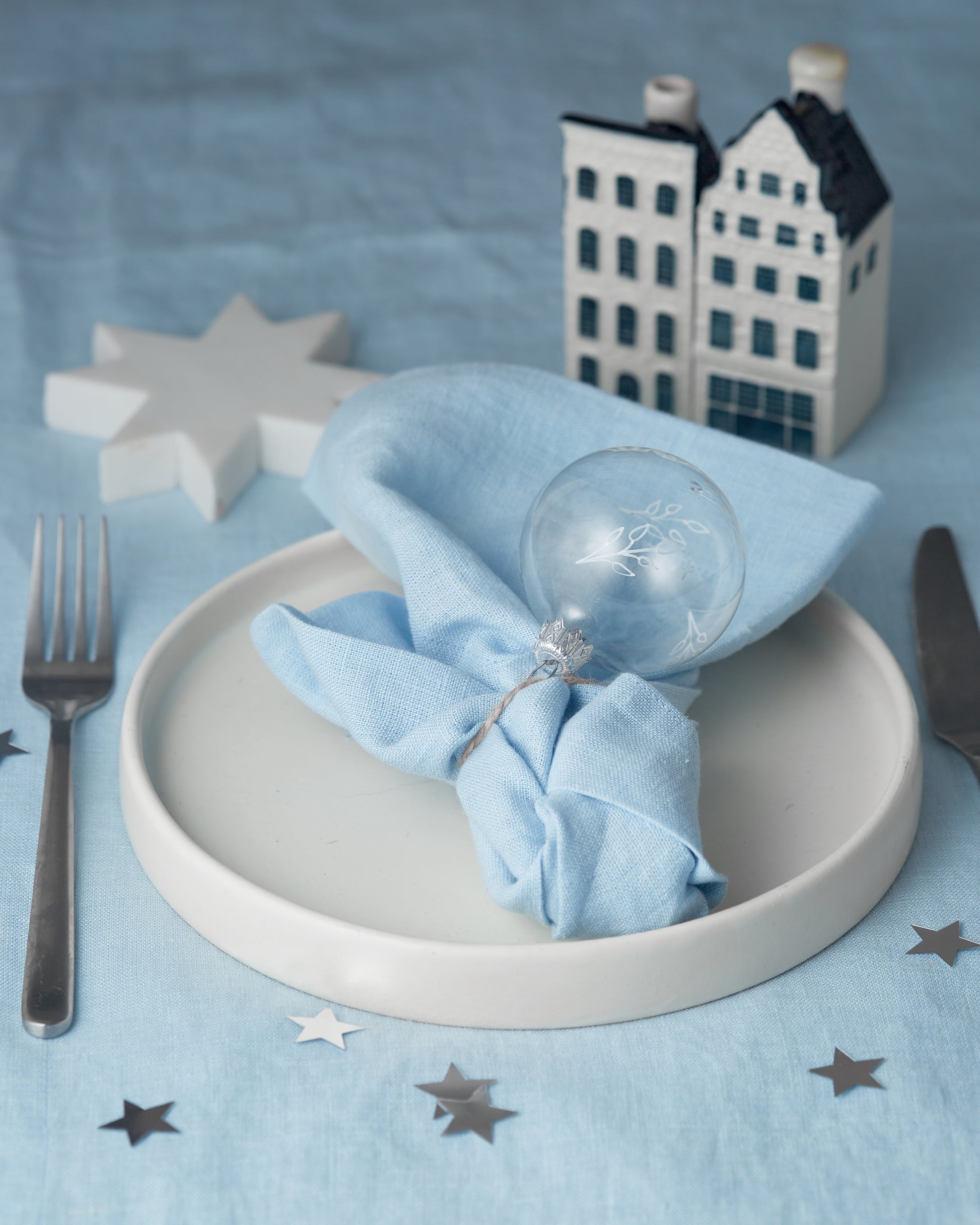 Table setting with a blue napkin folded around a glass ornament on a white plate, on a blue tablecloth with star decorations.