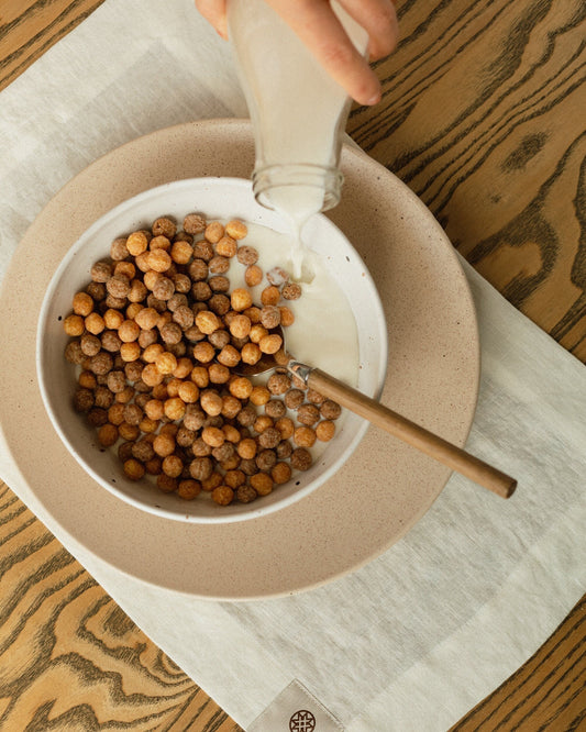 Hand pouring milk into a bowl of cereal on a linen placemat with wooden spoon